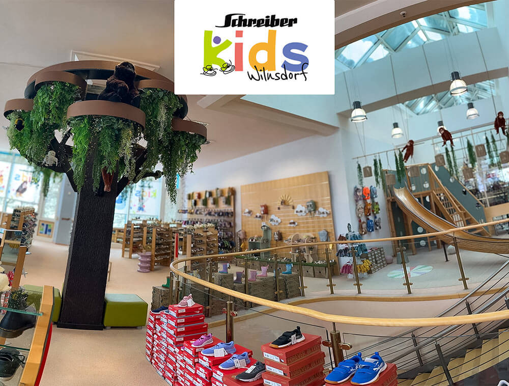 Innenansicht von Schreiber Kids Adventureland in
Wilnsdorf: großer Baum der Wünsche in der Mitte des Stores,
Rutsche und Kinderschuhregale im Kinderschuhfachgeschäft
Siegerland